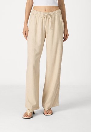 Pantalon beige ample avec taille à cordon, porté avec des sandales beiges à talons sur une personne debout sur un fond blanc.