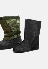 Dos pares de botas: uno en camuflaje verde con suela de goma y parte superior ajustable, el otro en fieltro gris oscuro con un acento de cuero negro.