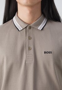 Muškarac nosi taupe majicu s kratkim rukavima s ovratnikom crne i bijele pruge te crnim izvezenim BOSS logotipom na lijevoj strani prsnog koša.