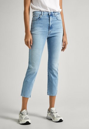 Persona vistiendo jeans cortos de color azul claro, camiseta blanca y zapatillas blanco-grises, de pie contra un fondo neutro.