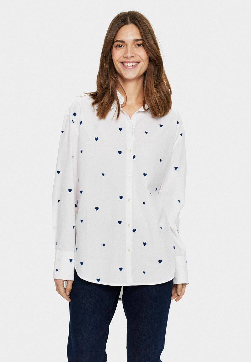 White button-up shirt featuring scattered navy blue heart patterns, with a classic collar and long sleeves. Worn with dark jeans.