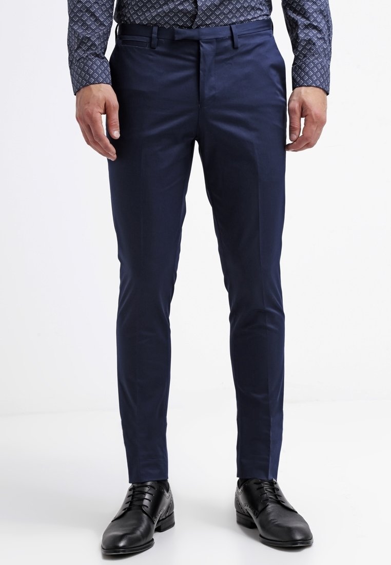 Homme portant un pantalon de costume bleu marine slim, des chaussures habillées en cuir noir et une chemise à manches longues à motifs, debout devant un fond blanc.