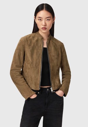 Nainen, jolla on suorat tummat hiukset, pukeutunut ruskeaan mokkanahkaiseen vetoketjulliseen takkiin mustan topin ja mustien housujen päällä, kädet taskuissa.