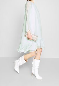 Robe vert clair, longueur genou, avec de longues manches et un ourlet froncé. Associée à des bottes blanches ample et une pochette argentée pailletée.