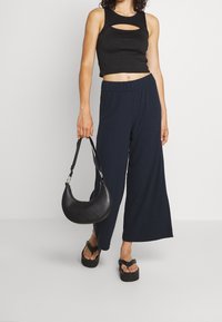Black ribbed crop top with cutout detail, paired with navy wide-leg pants. Holds a black leather crescent-shaped handbag; footwear features thick soles.