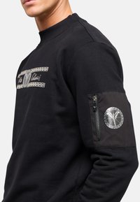 Sort sweatshirt med ribbet krave, der har et sølvlogo på venstre ærme og dekorativ tekst hen over brystet i et kontrasterende mønster.