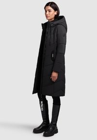 Manteau doudoune long noir avec capuche, présentant un matelassage en chevrons, une fermeture éclair à l'avant et des poches latérales. Associé avec des bottes noires à hauteur de cuisse.