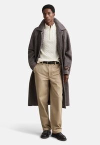 Brun ternet uldfrakke med stor krave, cremefarvet kabelstrikket sweater, beige bukser og sorte sko. Lige snit med løs pasform.