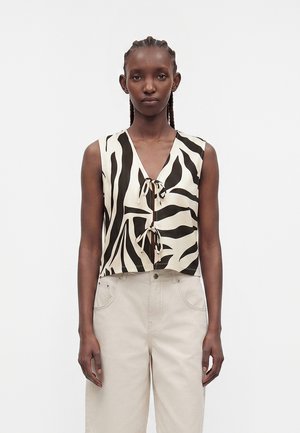 Γυναίκα με κοτσίδες που φοράει αμάνικη μαύρο-άσπρη κορυφή με ρίγες ζέβρας και μπροστινούς δεσίμους και ψηλόμεσο ανοιχτό μπεζ παντελόνι.