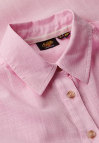 Camisa de color rosa claro con tejido texturizado, cuello abotonado y dos botones en la tapa. Presenta un detalle en el interior del collar en contraste.