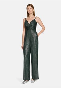 Combinaison métallique verte avec un profond décolleté en V, un corsage plissé et des jambes larges. Caractérisée par des bretelles fines et deux poches latérales. Escarpins argentés.