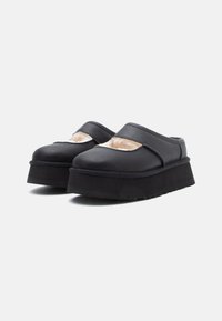 Sapatos pretos de plataforma, tipo slip-on, com solas grossas e forro de pelo bege visível através de recortes na parte superior, colocados sobre um fundo branco.