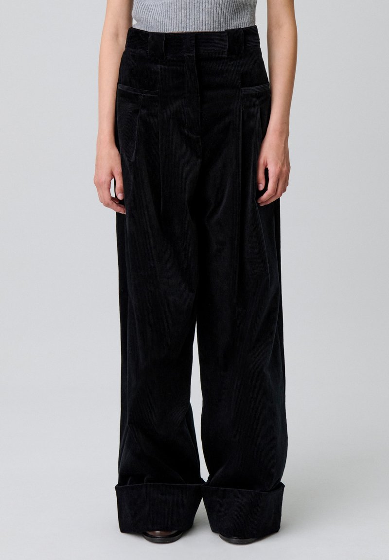 Pantalones de pana ancha de color negro con una cintura alta, dos bolsillos frontales y un dobladillo enrollado. Tejido texturizado con estrías verticales.