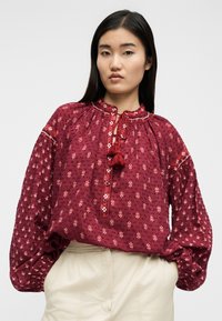 Femme portant un chemisier rouge à fleurs, ample et à manches longues, avec des glands au niveau de l'encolure, et un pantalon beige, debout les mains dans les poches.