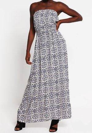 Vrouw draagt een strapless, witte jurk tot op de grond met zwarte geometrische en bloemmotieven, gecombineerd met zwarte open-toe hakken.