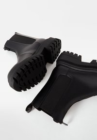 Černé gumové kotníkové boty s texturovanou podrážkou, elastickými bočními panely a poutky, s hladkým povrchem a robustním podpatkem.