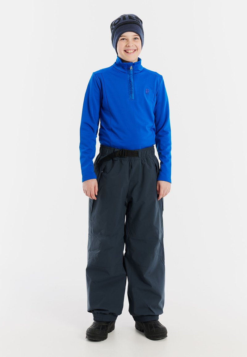 Blå långärmad tröja med dragkedja, kombinerad med löst sittande mörkgrå snöbyxor och svarta kängor. En halsduksliknande mössa fulländar outfiten.