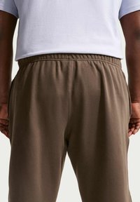 Pantalon de survêtement marron en tissu doux, avec une taille élastique et des poches latérales, vu de dos. T-shirt gris clair visible au-dessus.