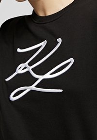 Camiseta negra con un destacado diseño blanco bordado con líneas fluidas, confeccionada en una tela suave y texturizada, que presenta un cuello redondo.