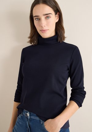 Navyblauwe turtleneck van ribbelstof, met lange mouwen en een relaxte pasvorm, gecombineerd met denim jeans en een neutrale achtergrond.