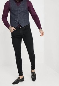 Gilet di lana grigia su una camicia bordeaux scuro, abbinato a jeans neri slim-fit e mocassini neri con un accento dorato.