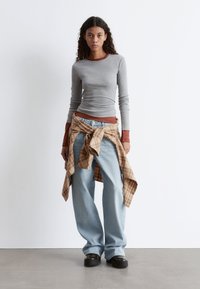 Graues Langarmshirt, das über einem braunen Top getragen wird, hellblaue weit geschnittene Jeans und ein kariertes Hemd um die Taille gebunden. Schwarze Plateauschuh.