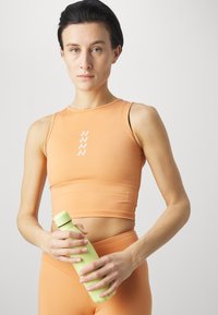 Sportovní crop top v broskvové barvě s přiléhavým střihem a bílým logem. Modelka drží světle zelenou lahvičku na vodu. Hladká tkanina, minimální švy.