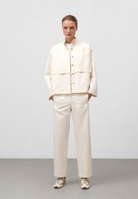 Giacca oversize color crema con bottoni frontali, tasca sul petto e maniche larghe, abbinata a pantaloni tapered coordinati e sneakers beige.