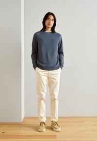 Blauer, strukturierten Pullover, crèmefarbene Hose und hellbraune Schnürschuhe. Stehende Pose vor einer neutralen Wand und einem Holzboden.