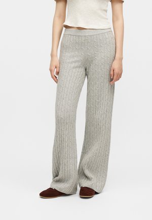 Žena má na sebe svetlosivé široké pletené nohavice s vrkočovým vzorom a krátky biely textúrovaný top, v kombinácii s tmavohnedými plochými topánkami.