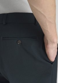 Ciemne spodnie z gładkiego materiału, z kieszeniami po bokach i z tyłu, z zapięciem na guzik w tylnej kieszeni oraz dopasowanym krojem.