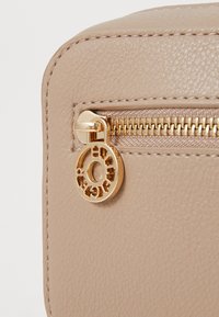 Coin de sac en cuir texturé beige avec une fermeture éclair dorée et un charme présentant un design découpé circulaire. Finition lisse et couture détaillée.