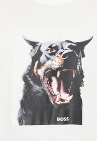 Melns un brūns grafisks attēls ar pīkstošu suni uz baltas T-krekla, attēlā redzamas asas zobu un zem attēla teksts "BOSS".