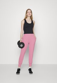 Juodas tank top'as, rožinės spalvos sportinės kelnės su kišenėmis ir juodi sportbačiai. Modelis laiko svorį. Paprastas, prigludęs dizainas, sklandus paviršius.