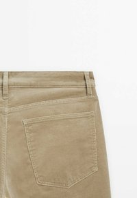 Pantalon en velours côtelé beige montrant la poche arrière et les passants de ceinture sur fond blanc, mettant en valeur la texture du tissu et les détails de la couture.
