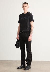 Sort bomulds t-shirt med "Calvin Klein" logo, parret med sorte jeans og sorte fodtøj. Holder en sort taske. Neutral baggrund.