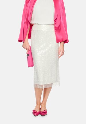 Frau, die einen weißen Pailletten-Midirock, einen knallpinkfarbenen Blazer und passende pinkfarbene spitze Ballerinas trägt, hält eine pinkfarbene Clutch in der linken Hand.