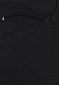 Sort denimstof med en glat tekstur, med en lomme og syningsdetaljer, inklusive en lille metalaccent nær lommen.