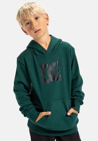 Zelená mikina s kapucí a velkým černým grafickým logem na přední straně, s klokaní kapsou a žebrovanými manžetami. Měkká, texturovaná tkanina.