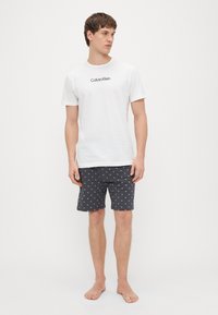 Bela bombažna majica s črnim logotipom "Calvin Klein", kombinirana s temnimi kratkimi hlačami z majhnim belim vzorcem sidra. Stoji bosa.