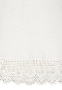 Weißer Stoff mit einem strukturierten Spitzenbesatz am Saum, der gewellte Kanten und dekorative Muster aufweist, darunter kleine Pompons und offene Webungen.