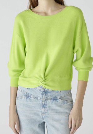 Kvinde iført limegrøn langærmet sweater snoet i taljen kombineret med lyseblå højtdækkende jeans, stående mod hvid baggrund.