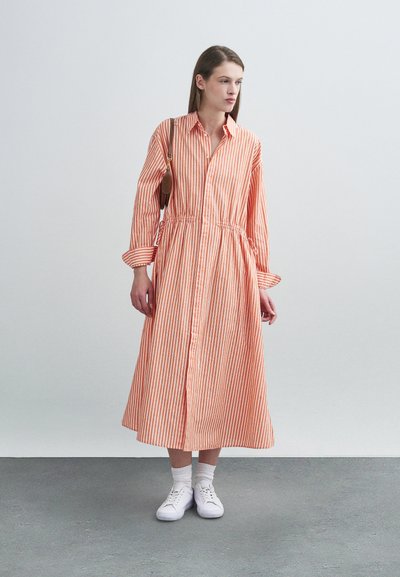 Φόρεμα με μακριά μανίκια και κουμπιά, σε πορτοκαλί και λευκές κάθετες ρίγες, εφαρμοστό στη μέση, φορεμένο με λευκά αθλητικά παπούτσια και κάλτσες.