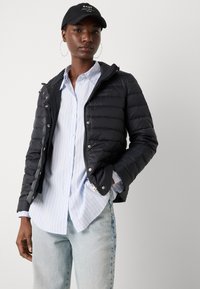 Black puffer jacket with a zip closure and snap buttons, worn over a light blue striped shirt. Features silver hardware and a fitted design.