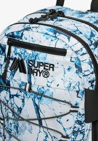 Niebieski i biały plecak z wzorem marmurowym, z wieloma kieszeniami zapinanymi na zamek oraz czarnymi elementami, z logo "Superdry".
