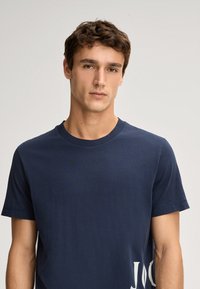 Marineblaue Baumwoll-T-Shirt mit Rundhalsausschnitt, kurzen Ärmeln und einem großen weißen Grafikdruck auf der unteren linken Seite. Glatte Textur.