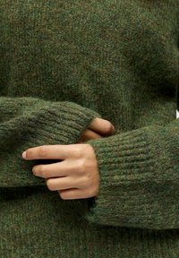 Grüner Strickpullover mit einer strukturierten, flauschigen Optik und gerippten Bündchen. Der Stoff weist eine Mischung aus dunklen und hellen Grüntönen auf.