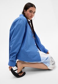 Blue checked shirt with long sleeves, buttoned cuffs, and a loose fit. Model wears a multi-colored skirt and black sandals, with braided hair.