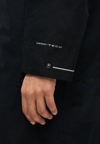 Χέρι με μέτριο χρωματικό τόνο δίπλα σε μαύρο μανίκι με μακρύ μανσέτα με ετικέτα "OMNI-TECH", μεταλλικό κουμπί ασφαλείας και αντανακλαστική λωρίδα.