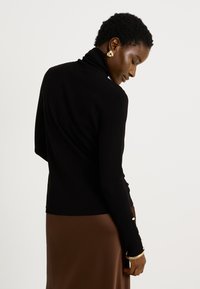 Jersey de cuello alto negro con mangas largas, textura acanalada y detalle decorativo de botones dorados en los puños. Tejido suave, diseño ajustado.